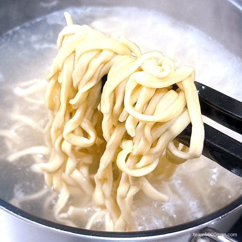 豚星。汁無し蕎麦 茹でている平打ち麺をトングで持ち上げたところ